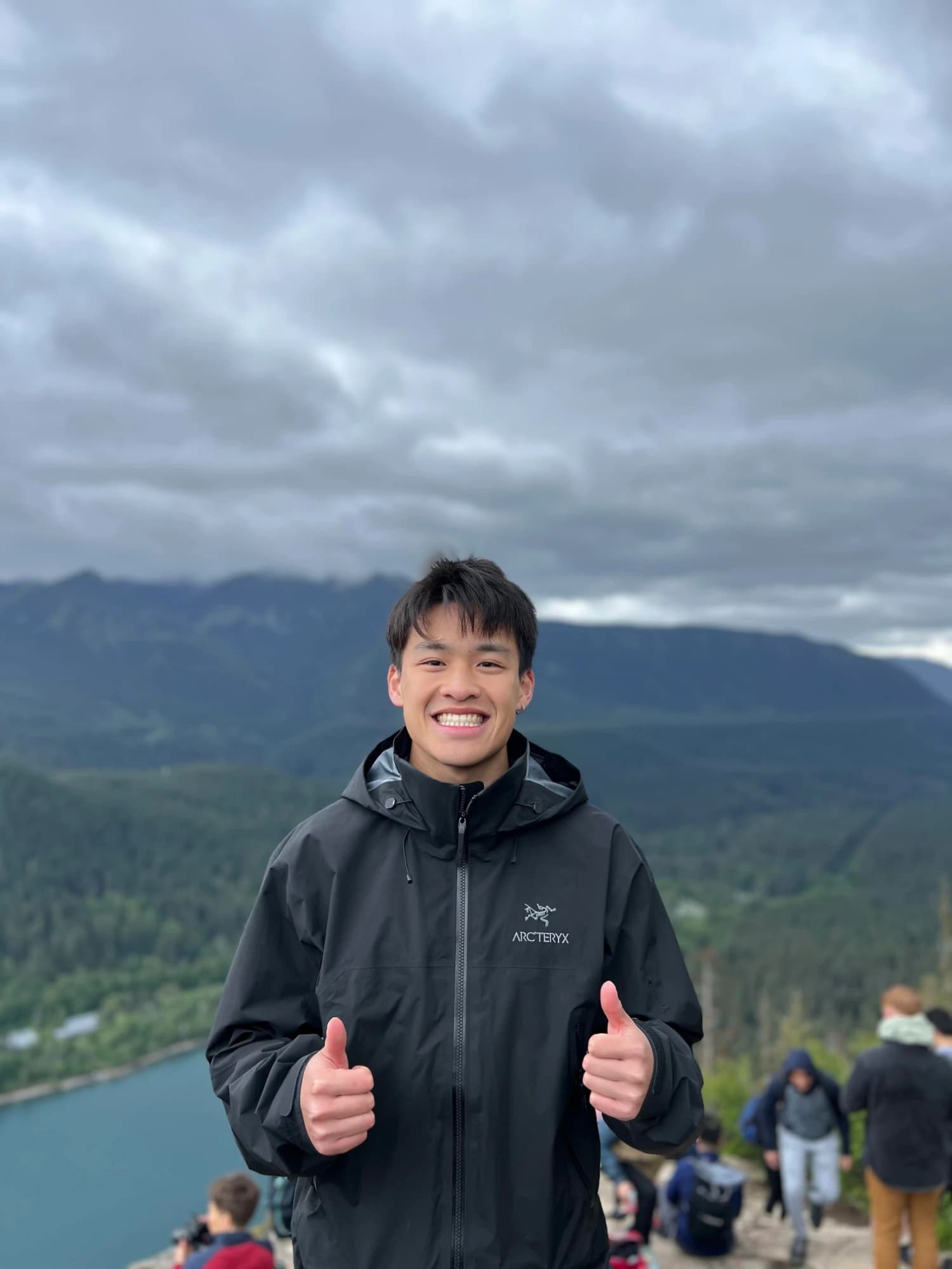 Brandon smiling with two thumbs up on a cloudy mountain summit, a lake and forested valley behind him.